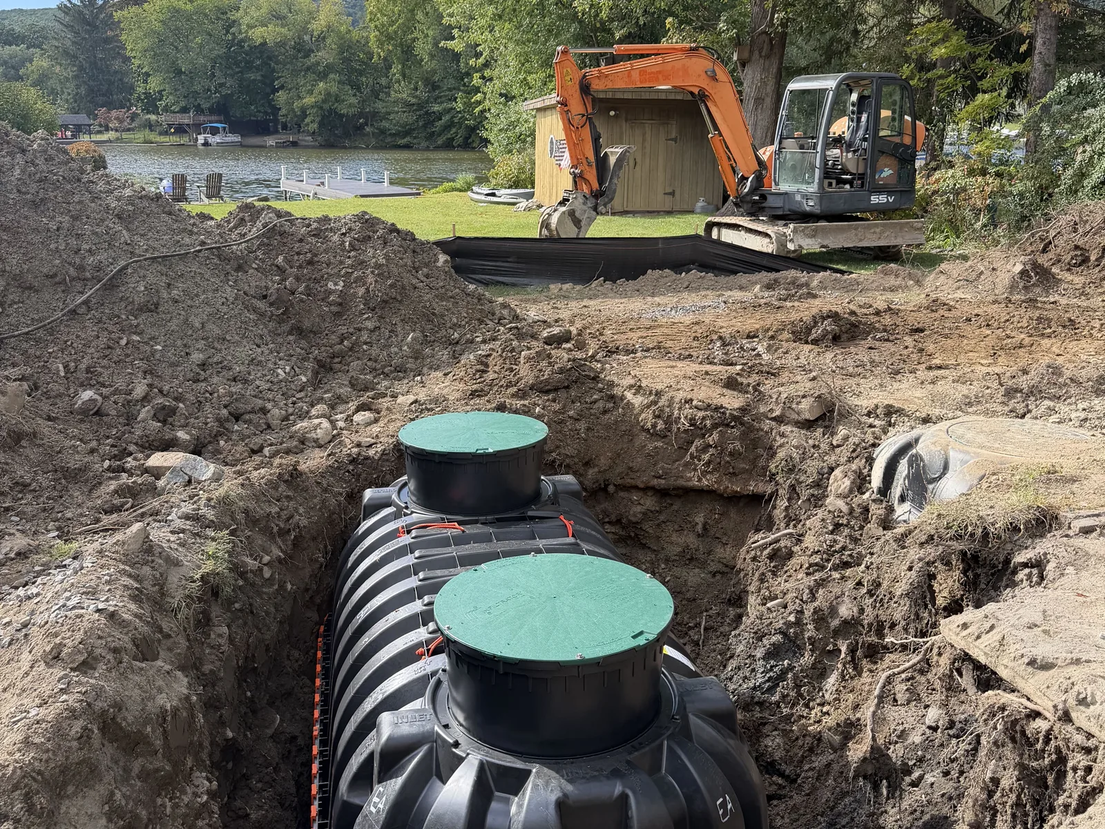 Our excavator beside the open tank hole on a lakefront lot, with the lake and docks behind