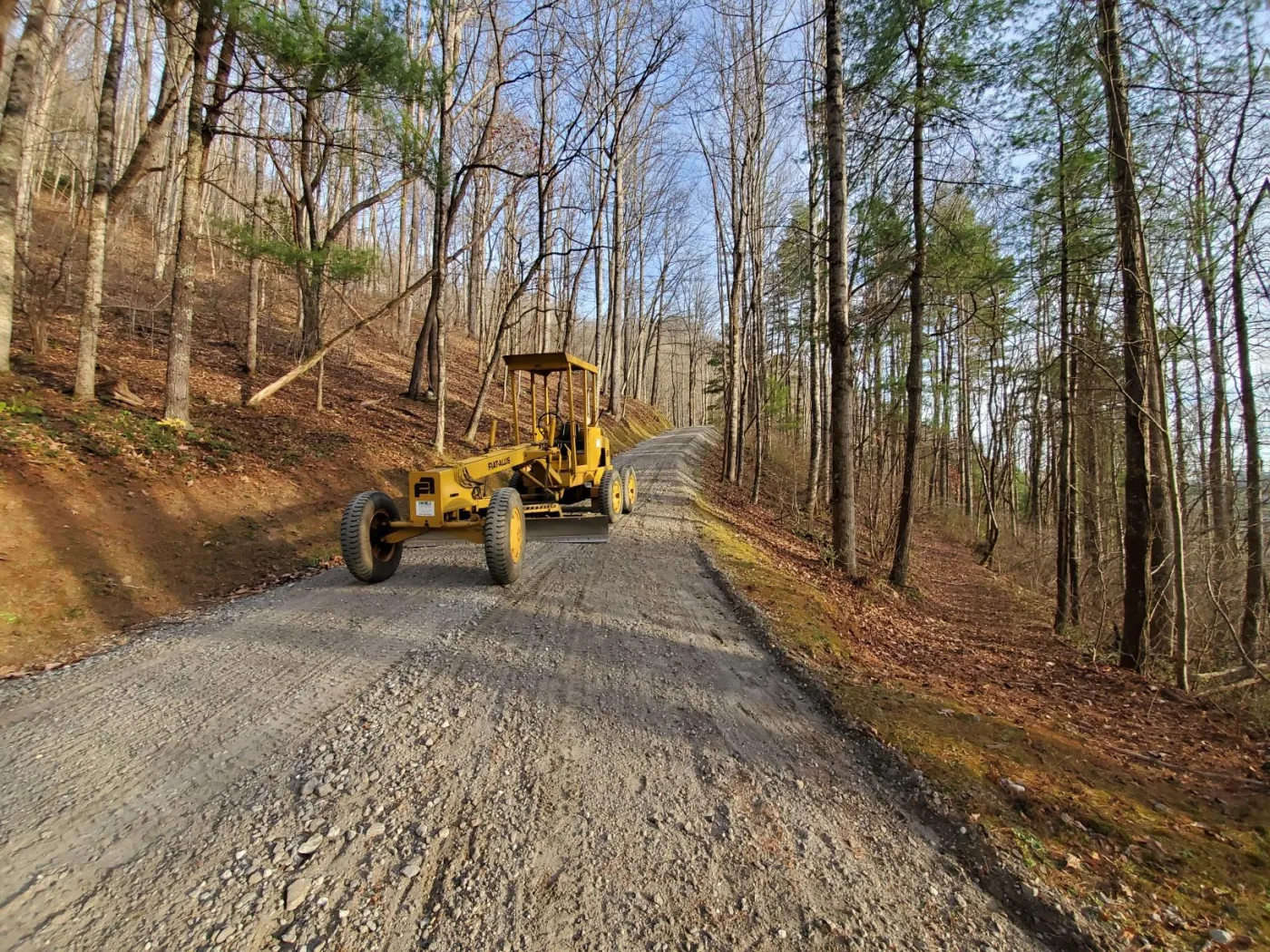 Motor grader shaping a freshly graded gravel lane climbing through bare hardwoods and pines