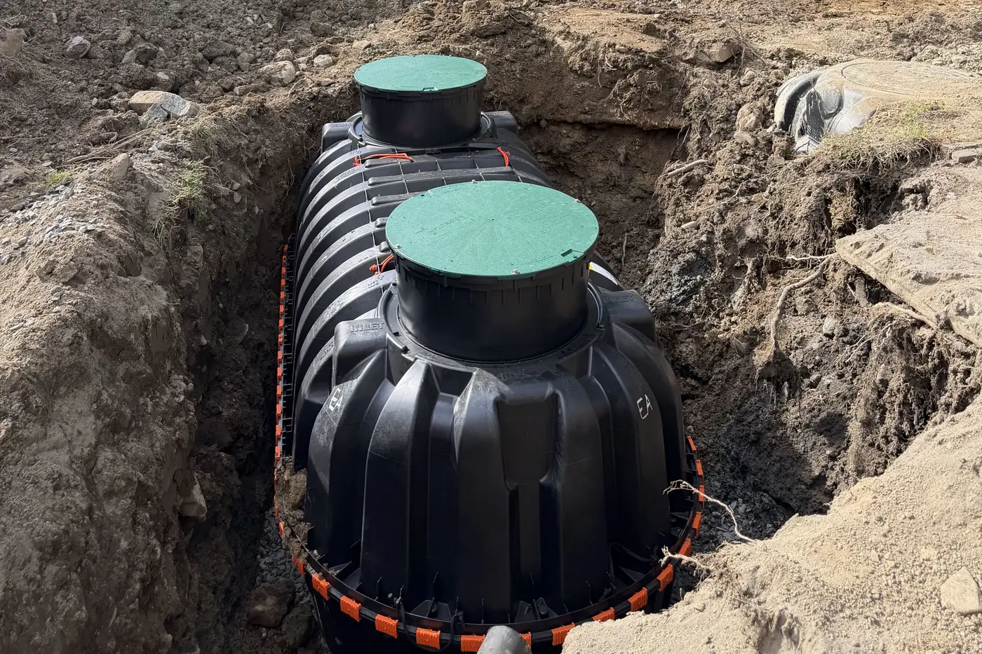 Black ribbed plastic septic tank with two green access lids set in a rocky excavation