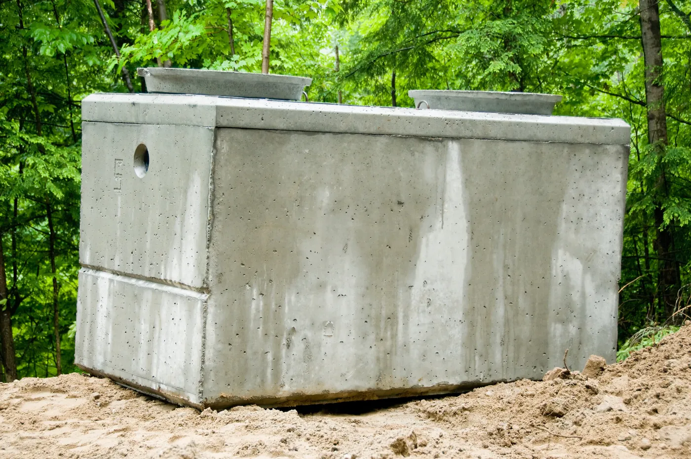 Precast concrete septic tank with two lids set on sand at the edge of green woods