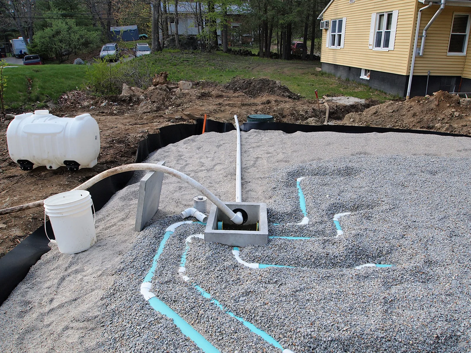 Stone bed with white laterals, a concrete distribution box and a plastic pump tank beside a house